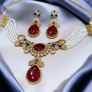May include: A multi-strand pearl necklace and matching drop earrings set featuring gold-tone metal, clear rhinestones, and large deep red teardrop-shaped stones. The necklace centerpiece includes colorful accent stones in blue, green, and yellow.