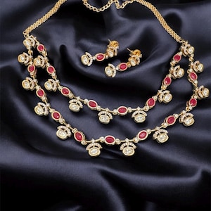May include: A gold-toned jewelry set featuring a necklace and matching earrings. The necklace has two strands with red oval-shaped stones and clear, round accents. The earrings have a similar design. The set is displayed on a dark, silky fabric.
