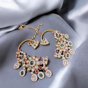 May include: Gold-toned earrings with a floral design, featuring a combination of clear, red, and green gemstones. The earrings are set against a gray satin background, showcasing their intricate details and craftsmanship.