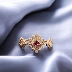 May include: A gold ring featuring a central square-cut red gemstone surrounded by a cluster of small round clear stones in a diamond-shaped setting, with two additional diamond-shaped side accents containing clear stones on the band.