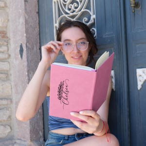 May include: A pink journal with a black floral design and the name "Chelsea" written in cursive. The journal is open and being held by a person wearing glasses.