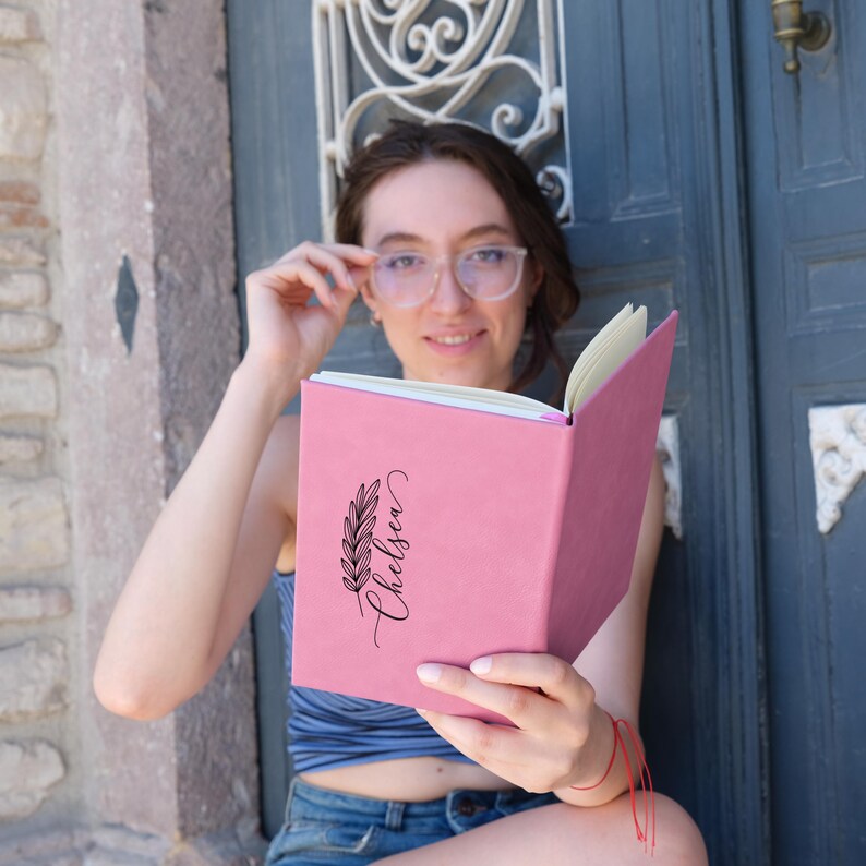 May include: A pink journal with a black floral design and the name "Chelsea" written in cursive. The journal is open and being held by a person wearing glasses.