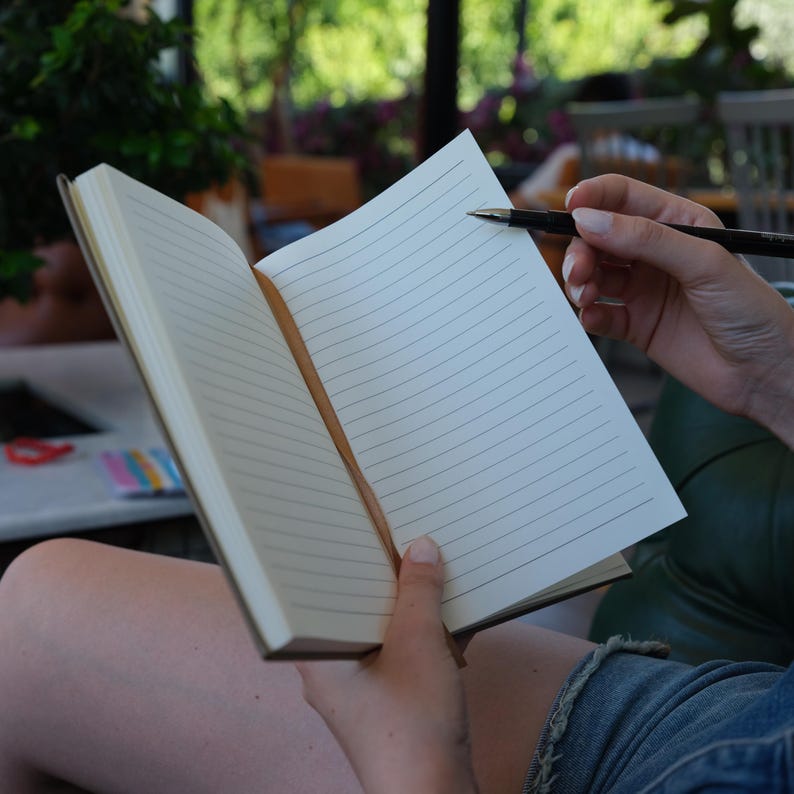 May include: A blank lined notebook with a brown leather cover is open and being held by a person. A pen is resting on the open pages.