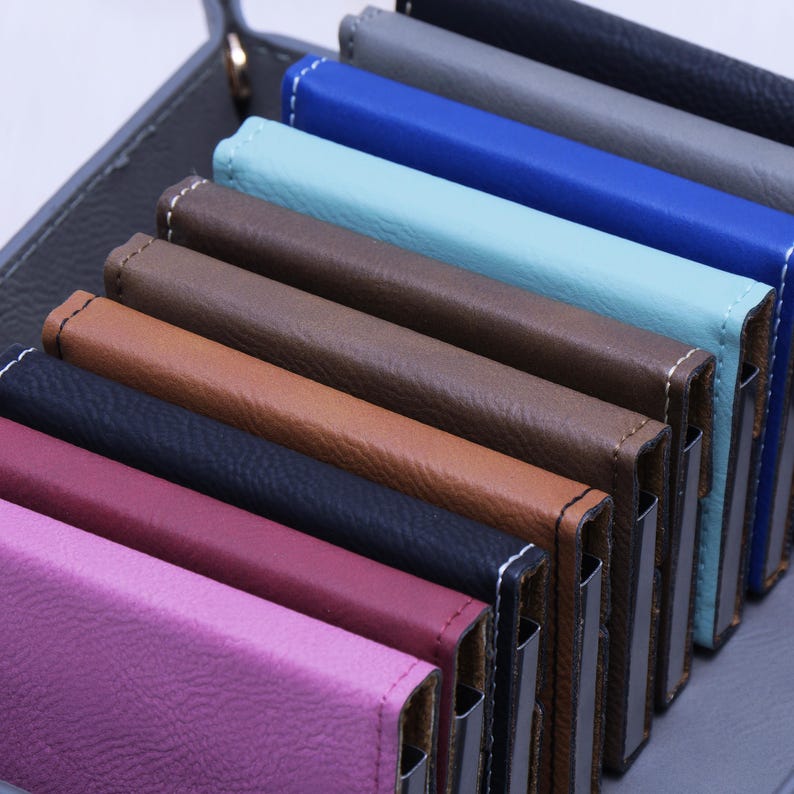 K&ouml;nnte beinhalten: Ein Set aus zehn Lederkartenetuis in verschiedenen Farben, darunter rosa, rot, schwarz, braun, beige, blau, t&uuml;rkis und grau. Die Etuis sind &uuml;bereinander gestapelt, wobei das oberste Etui rosa ist.