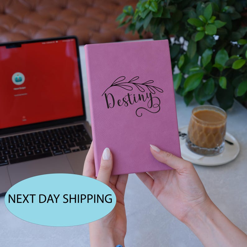 May include: A pink leather-bound journal with the word "Destiny" written in black script on the front cover. The journal has a floral design on the front cover.