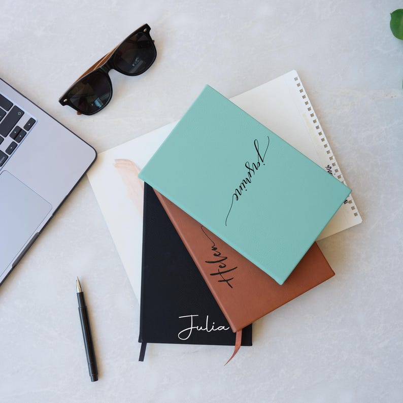 May include: Three notebooks stacked on top of each other. The top notebook is teal and says "unrushed!" in black cursive. The middle notebook is brown and says "Helen" in black cursive. The bottom notebook is black and says "Julia" in white cursive.