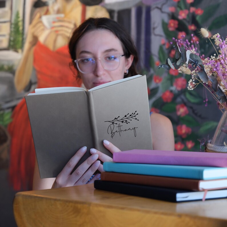 May include: A brown leather-bound journal with the name "Bethany" and a small floral design on the front cover. The journal is open and being read by a person.