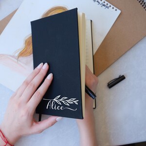 May include: A black leather-bound journal with a white floral design and the name "Alice" written in cursive on the front cover.