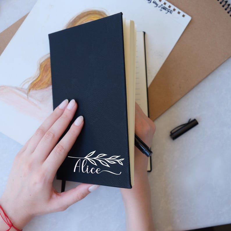 May include: A black leather-bound journal with a white floral design and the name "Alice" written in cursive on the front cover.