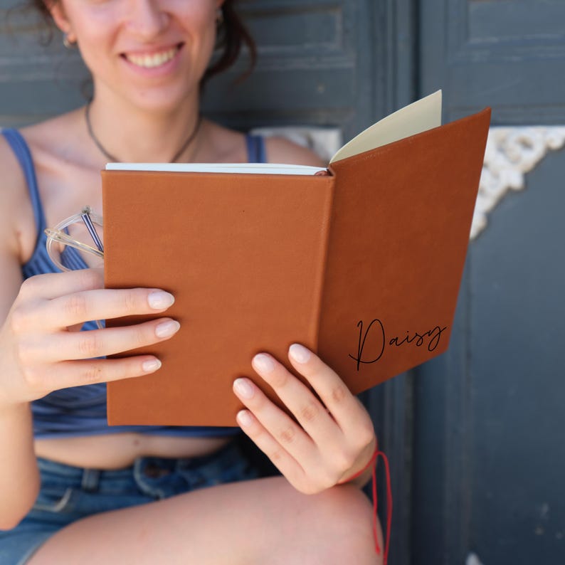 May include: A brown leather-bound journal with the name "Daisy" written on the cover. The journal is open and being held by a person.