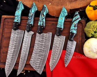 May include: A collection of six kitchen knives with patterned blades and turquoise and black handles. The blades have a textured, hammered appearance. The handles feature a marbled turquoise and black design, with gold-coloured rivets. The knives are displayed on a wooden chopping board.