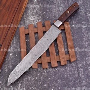 Puede incluir: Un cuchillo de cocina largo, dentado, de acero inoxidable con mango de madera. El cuchillo tiene un patrón de acero damasco y está apoyado sobre una superficie de listones de madera.
