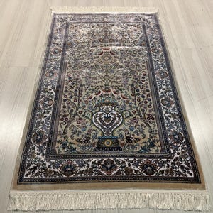 May include: A rectangular beige rug with an intricate floral pattern. The rug showcases a central vase design, encircled by detailed flowers and foliage in blue, green, and brown. The border is a darker shade with a fringe.