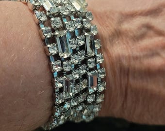 May include: A silver-toned bracelet with multiple rows of clear, sparkling rhinestones. The bracelet features a mix of rectangular and square-cut stones, creating a dazzling effect. The jewelry is designed to be worn around the wrist.