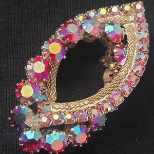 Puede incluir: Broche vintage dorado con forma de hoja abstracta, adornado con pedrería iridiscente multicolor. El broche presenta un marco dorado en forma de malla y está sobre un fondo oscuro.