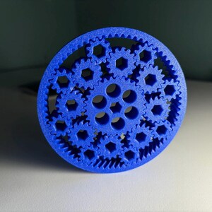 Può includere: Un meccanismo a ingranaggi blu stampato in 3D con più ingranaggi interconnessi. L'ingranaggio centrale ha una serie di ingranaggi più piccoli che lo circondano, il tutto all'interno di un telaio circolare. Gli ingranaggi hanno un design dettagliato e intricato.