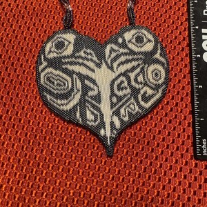 May include: A black and white beaded heart-shaped pendant necklace with a geometric design. The necklace is on a red background.