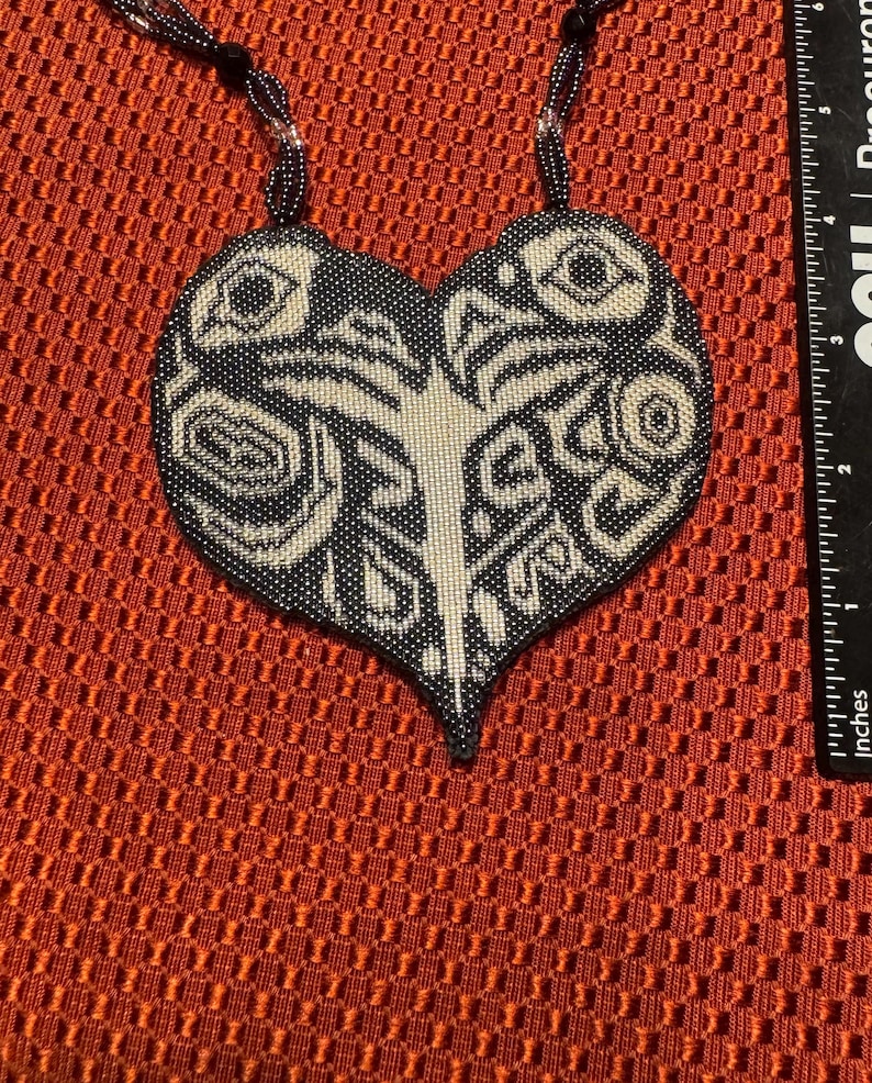 May include: A black and white beaded heart-shaped pendant necklace with a geometric design. The necklace is on a red background.