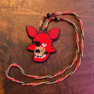 May include: A beaded necklace featuring a red fox head pendant with a black eye patch and white teeth. The pendant is red, black, and tan. The necklace cord is red, black, and gold. The necklace is on a wooden surface.