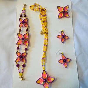 May include: A set of beaded jewelry featuring yellow and purple flower-shaped charms. The set includes a necklace, bracelet, and earrings.
