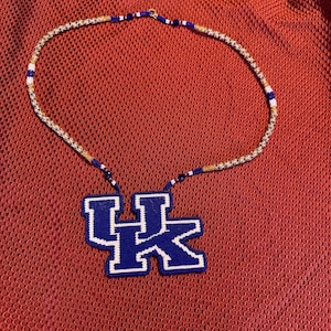 May include: A beaded necklace with a blue and white UK logo pendant. The necklace is made with white, gold, and blue beads.
