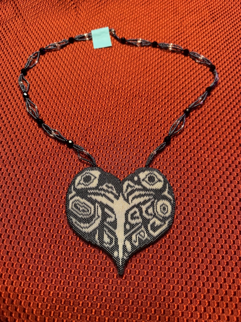 May include: A black and white beaded necklace with a heart-shaped pendant. The pendant features a geometric design with intricate details.