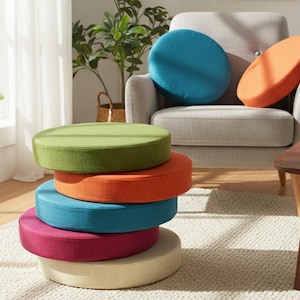 May include: A stack of six round floor cushions in various colors: green, orange, blue, magenta, and cream. Two additional cushions in blue and orange rest on a gray armchair. The cushions are on a cream-colored rug in a living room setting.