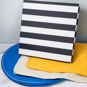 May include: A stack of three fabric cushions. The top cushion is black and white striped, the middle cushion is yellow, and the bottom cushion is cream. The cushions are sitting on a blue tray.