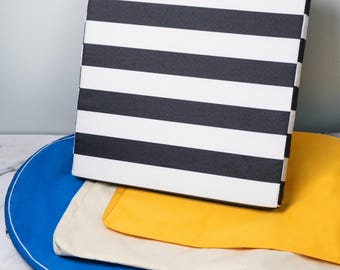 May include: A stack of three fabric cushions. The top cushion is black and white striped, the middle cushion is yellow, and the bottom cushion is cream. The cushions are sitting on a blue tray.