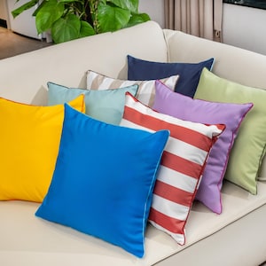 May include: A collection of square throw pillows in various colors, including yellow, blue, green, and purple. Some pillows feature striped patterns, while others are solid. The pillows are arranged on a cream-colored sofa.