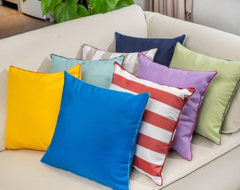May include: A selection of square cushions in various colours, including yellow, blue, green, and purple. Some cushions feature striped patterns, while others are solid. The cushions are arranged on a cream-coloured sofa.
