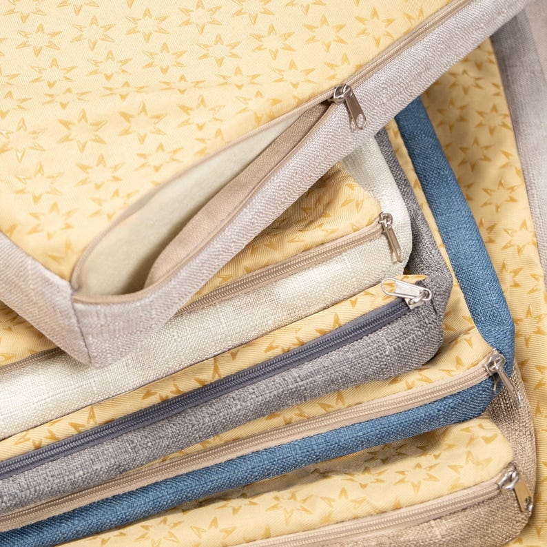 May include: A stack of zippered pouches in various shades of beige, grey, and blue. The pouches are made of a soft, textured fabric and have a star pattern.