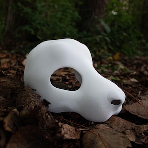 Könnte beinhalten: Eine glatte, matte weiße Tiermaske mit einer abgerundeten Schnauze und einer einzelnen großen, ovalen Augenöffnung. Die Maske hat eine konturierte, skulpturale Form mit einer definierten Nasenspitze und einem geschwungenen Profil.