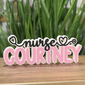 May include: Pink and white wooden sign with the text "nurse COURTNEY" and a black stethoscope with a heart on the end.