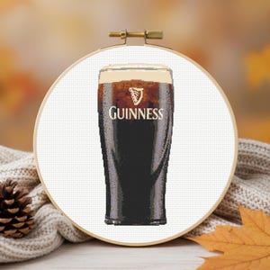 Pu&ograve; includere: Un telaio da ricamo a punto croce con una pinta di Guinness. La stout scura &egrave; sormontata da una testa cremosa e il logo Guinness &egrave; visibile. Lo sfondo include un maglione accogliente, una pigna e foglie autunnali.
