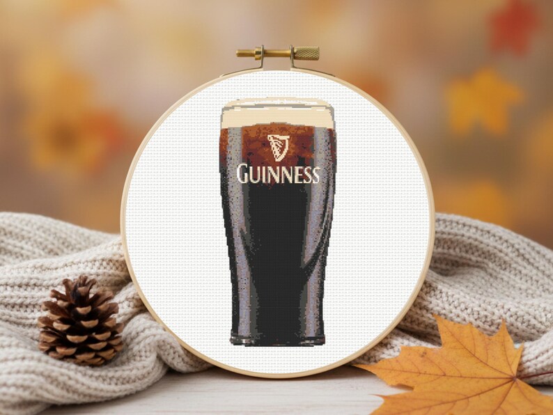 Pu&ograve; includere: Un telaio da ricamo a punto croce con una pinta di Guinness. La stout scura &egrave; sormontata da una testa cremosa e il logo Guinness &egrave; visibile. Lo sfondo include un maglione accogliente, una pigna e foglie autunnali.