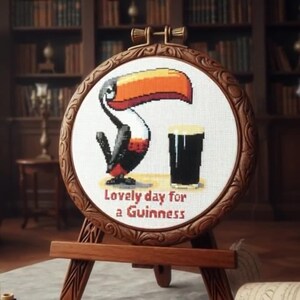 May include: A cross-stitch embroidery hoop featuring a toucan and a pint of Guinness. The toucan has a large orange and black beak, with black and red feathers. The text "Lovely day for a Guinness" is below the image. The hoop is displayed on a wooden easel.