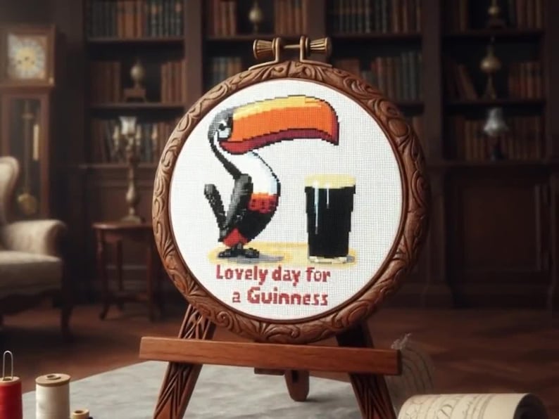May include: A cross-stitch embroidery hoop featuring a toucan and a pint of Guinness. The toucan has a large orange and black beak, with black and red feathers. The text "Lovely day for a Guinness" is below the image. The hoop is displayed on a wooden easel.