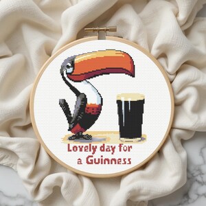 May include: Cross-stitch embroidery in a wooden hoop features a toucan with a large orange and red beak, a pint of Guinness, and the text "Lovely day for a Guinness." The design is set against a white background.
