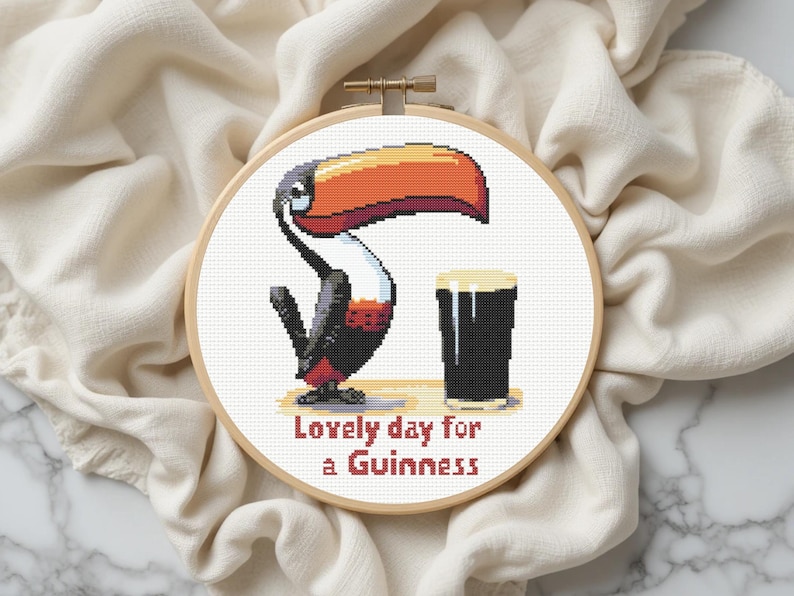 May include: Cross-stitch embroidery in a wooden hoop features a toucan with a large orange and red beak, a pint of Guinness, and the text "Lovely day for a Guinness." The design is set against a white background.
