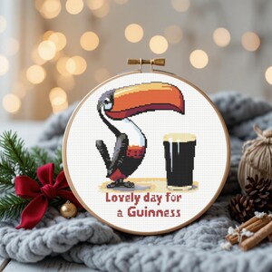 May include: Cross-stitch hoop art featuring a toucan with a large orange and yellow beak, a black body, and red accents. The toucan is next to a pint of dark beer. Text reads "Lovely day for a Guinness."