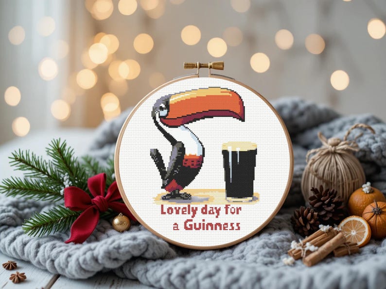 May include: Cross-stitch hoop art featuring a toucan with a large orange and yellow beak, a black body, and red accents. The toucan is next to a pint of dark beer. Text reads "Lovely day for a Guinness."