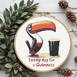 May include: A cross-stitch embroidery hoop featuring a pixelated toucan with an orange beak and a pint of Guinness. The text "Lovely day for a Guinness" is stitched below the image. The hoop is surrounded by greenery and pinecones.