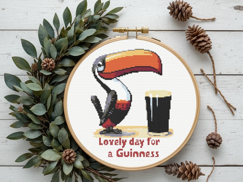 May include: A cross-stitch embroidery hoop featuring a pixelated toucan with an orange beak and a pint of Guinness. The text "Lovely day for a Guinness" is stitched below the image. The hoop is surrounded by greenery and pinecones.