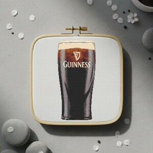 Pu&ograve; includere: Un punto croce di una pinta di Guinness stout in un bicchiere, con il logo e il testo Guinness. L'immagine &egrave; incorniciata in un telaio da ricamo quadrato in legno. Lo sfondo &egrave; una superficie grigio chiaro con elementi decorativi.