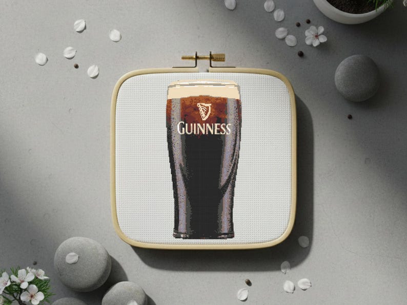 Pu&ograve; includere: Un punto croce di una pinta di Guinness stout in un bicchiere, con il logo e il testo Guinness. L'immagine &egrave; incorniciata in un telaio da ricamo quadrato in legno. Lo sfondo &egrave; una superficie grigio chiaro con elementi decorativi.