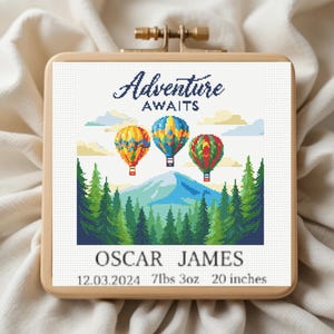 May include: Cross-stitch artwork in a wooden hoop features three colorful hot air balloons above a mountain range and evergreen trees. The text "Adventure Awaits" is above the balloons. Below are the names "OSCAR JAMES", date, weight, and 20 inches.