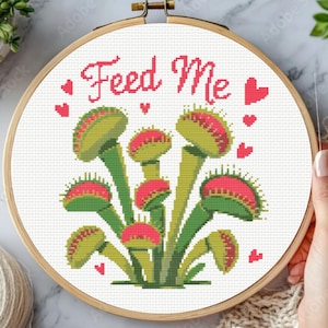 May include: Cross-stitch embroidery in a wooden hoop features a Venus flytrap design with the words "Feed Me" in red. The flytraps are green and red, with small red hearts scattered around the design. A needle is visible.
