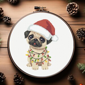 May include: A cross-stitch design of a pug wearing a Santa hat and a string of Christmas lights. The pug is tan, black, and white, with a red and white Santa hat. The design is set in a round embroidery hoop.