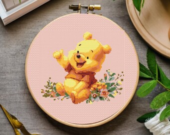 May include: A cross-stitch embroidery hoop with a pink background featuring a smiling yellow cartoon bear with brown eyes and a brown shirt. The bear is surrounded by colorful flowers.